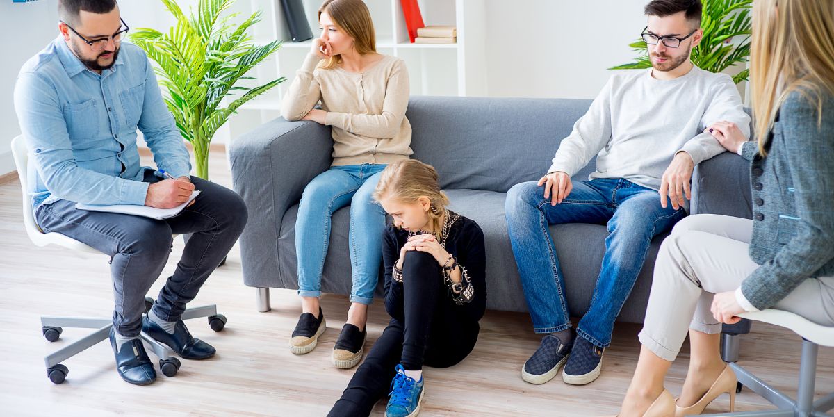 Therapist taking notes while parents sit on a couch and a young girl sits on the floor, appearing upset, during a family therapy session