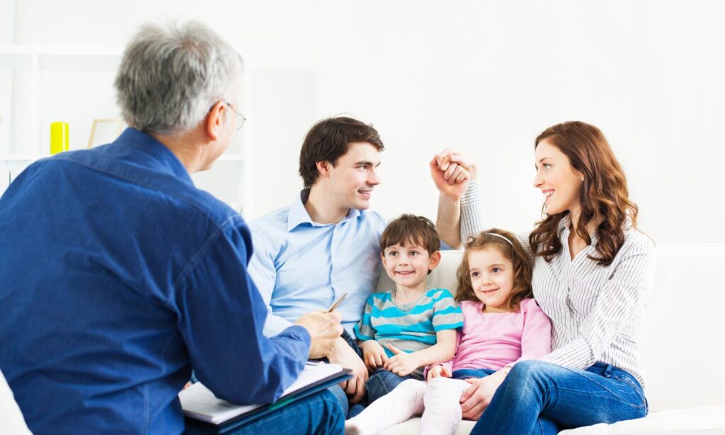 Parents and two young children smiling and holding hands, speaking to a male therapist during a family counseling session.