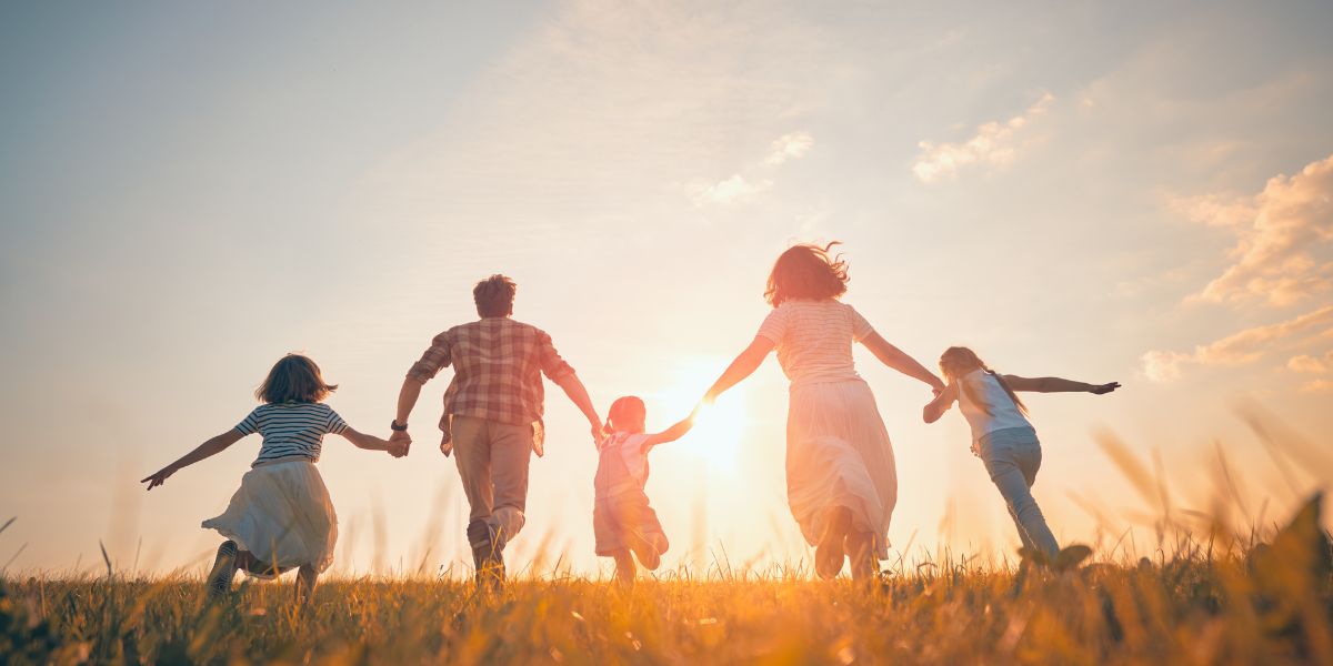 Family of five holding hands and running through a field at sunset, with bright sunlight in the background.